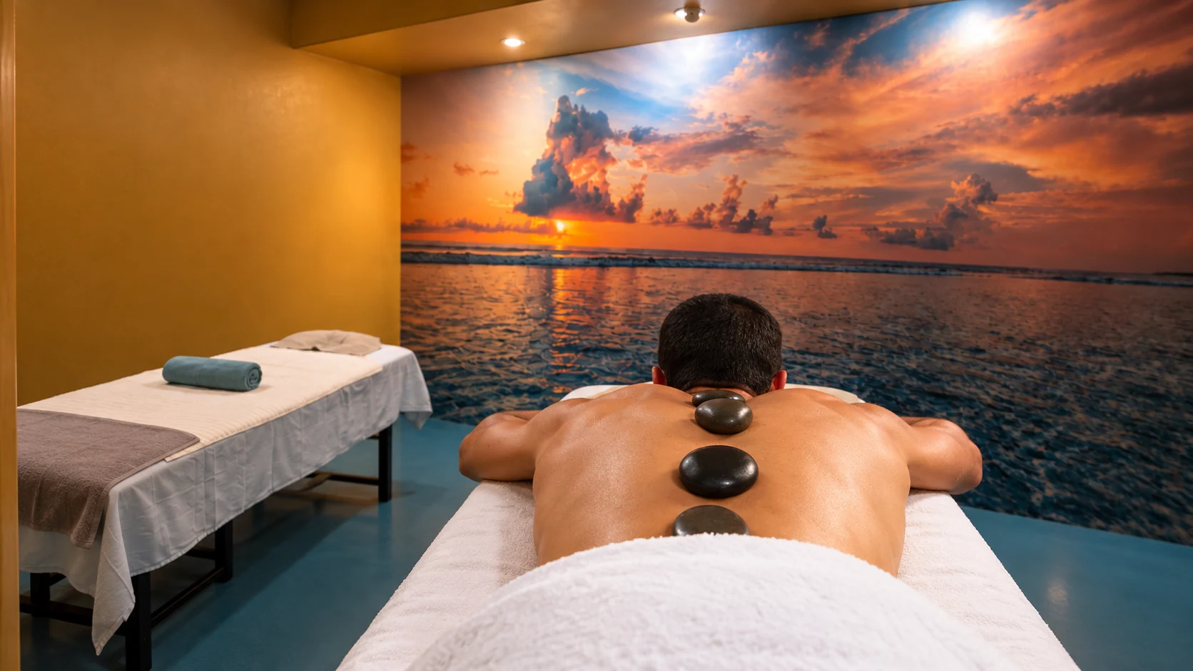 Terapia con Piedras Calientes Volcánicas en Tijuana – Masaje Milagroso Spa Masaje con piedras calientes volcánicas sobre la espalda en cabina con mural del océano al atardecer en Masaje Milagroso Tijuana